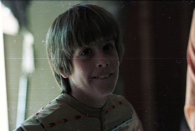 Vintage-style close-up of a young boy with short, parted hair, smiling slightly. He wears a floral-patterned shirt with red a...