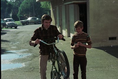 Two boys in 1960s-70s streetwear—striped sweater, button-up shirt, and trousers—pose with a classic steel-framed bicycle. Urb...