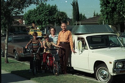 Family portrait in a suburban driveway, 1970s USA. Three children on bicycles—one with training wheels—pose beside an older m...