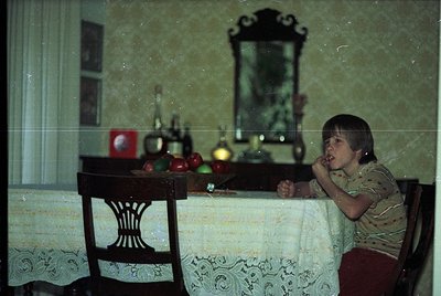 Vintage indoor scene featuring a child seated at a wooden table draped with a lace tablecloth. Reflective mirror reveals a fr...