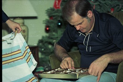 Vintage holiday scene featuring a man assembling a Scrabble game on a striped tablecloth, likely mid-1970s. Decor includes a ...
