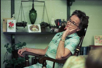 Mid-century woman in floral blouse poses thoughtfully in vintage armchair, surrounded by retro decor: scales, framed botanica...