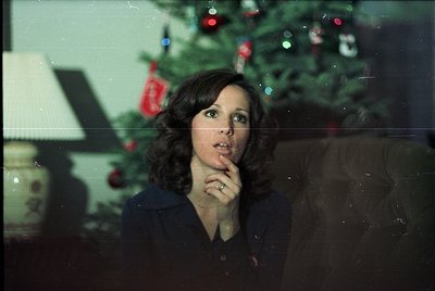Mid-century woman in a dark blouse poses thoughtfully indoors beside a decorated Christmas tree with red baubles and tinsel, ...