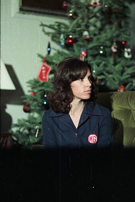 A woman in a dark blue jacket with a red "MG" logo poses beside a decorated Christmas tree, likely from the 1960s–1970s. The ...