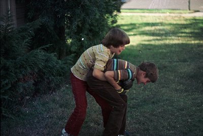 Two boys in 1970s-style clothing play outdoors in a residential yard. The boy on left wears a striped sweater and maroon pant...