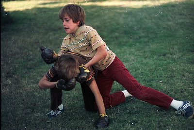 Two boys wrestling on grassy field, 1970s-era clothing: striped polo shirt, maroon pants, white socks with stripes. Candid ch...