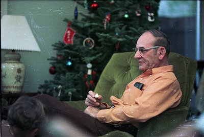 Mid-century man in retro glasses and orange sweater relaxes indoors beside a decorated Christmas tree with red/white ornament...