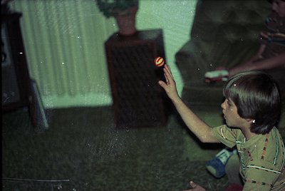Vintage indoor scene featuring a child reaching toward a glowing orange lightbulb in a dark room. Green-tinted lighting and p...