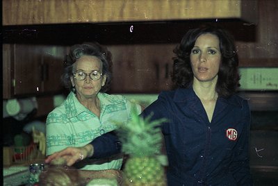 Vintage indoor portrait featuring two women in a kitchen setting, likely . Elderly woman in patterned blouse sits beside youn...