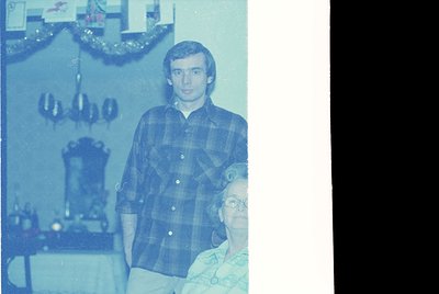 Vintage sepia-toned indoor portrait from the 1970s–1980s, featuring a man in a plaid shirt standing beside an elderly woman i...