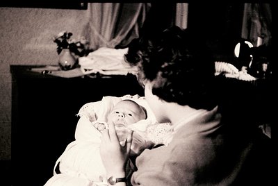 A tender black-and-white moment: a woman in a lace dress cradles a swaddled infant indoors, likely post-1950s. Soft lighting ...