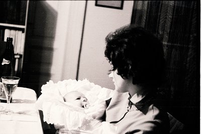 A tender black-and-white moment: a woman cradles a newborn wrapped in a lace-trimmed swaddle, seated at a table with a champa...