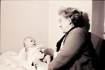 A tender black-and-white moment of a woman in mid-20th-century glasses and a cardigan cradling an infant in a simple room. Th...