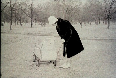 Black-and-white winter scene showing a woman in mid-1900s attire pushing a manual wheelbarrow filled with snow in a park. She...
