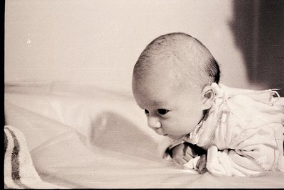 Black-and-white portrait of an infant crawling on a soft, textured surface, possibly a blanket. The child’s expression and po...