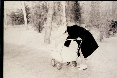 Mid-20th century black-and-white photo of a person pushing a vintage stroller on a snow-covered path. Hooded coat and white s...