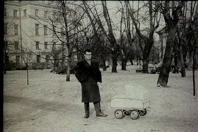 Mid-20th century urban park scene: Man in overcoat and cap pushes a small, wheeled laundry cart through snow-covered ground. ...