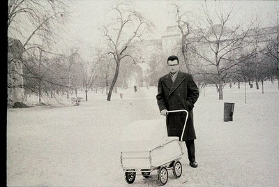 Mid-20th century man in winter coat pushes a wooden wheelbarrow on snow-covered park path, flanked by bare trees. Classic urb...