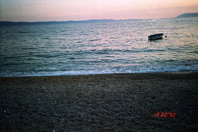 Vintage seaside scene with a small wooden boat anchored near shore at dusk. Waves gently lap against the pebbled beach under ...