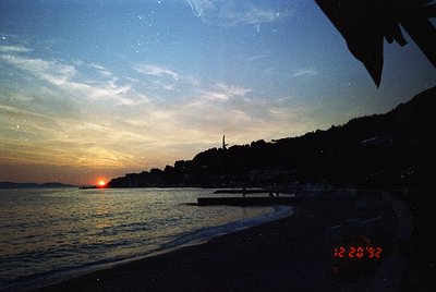 Sunset silhouette over coastal town with illuminated skyline. Low-angle shot captures waterfront buildings, a lone boat, and ...