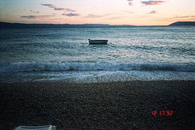 Golden-hour seascape with gentle waves lapping a pebbled shore. A lone wooden boat rests anchored near the horizon, silhouett...