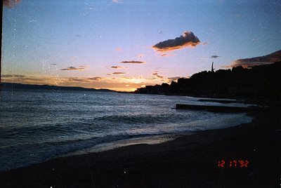 Vintage seaside sunset with golden horizon and scattered clouds over calm waters. Silhouetted trees and distant structures fr...