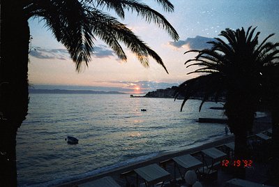 Sunset over a coastal landscape with palm trees framing the view. The horizon shows a distant shoreline with silhouetted stru...