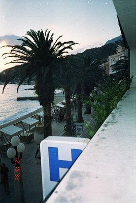 Seaside promenade lined with palm trees and modern Mediterranean-style buildings. Prominent blue "H" sign suggests hotel or h...