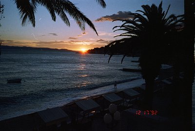 Sunset over a calm coastal scene, framed by tropical palm fronds. The digital timestamp (12:17:38) suggests late evening. Emp...
