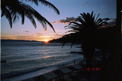 Sunset over a coastal horizon framed by tropical palm fronds, captured 12/17/92. Warm golden light reflects on calm waters, w...
