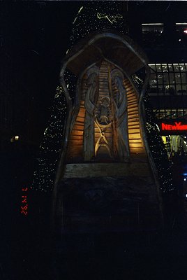 Relief sculpture of a robed figure holding a cross, illuminated at night in an urban setting. The carving features intricate ...
