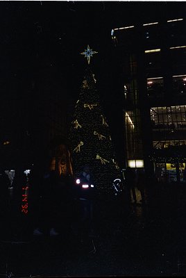 A towering, illuminated Christmas tree adorned with golden ornaments and a star-topped crown stands in an urban plaza. Neon s...