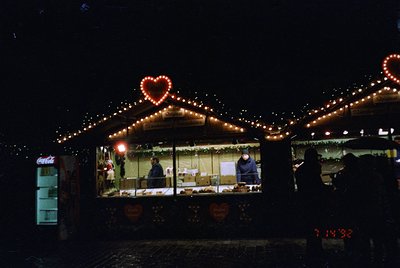 Nighttime market stall adorned with warm string lights and heart-shaped decorations. Indoor seating with blurred figures, lik...
