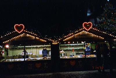Christmas market stalls adorned with heart-shaped string lights, illuminated at night. Vendors behind counters display baked ...