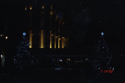 Neon-lit industrial facade at night, featuring four vertical cylindrical tanks with illuminated yellow-orange outlines. Refle...