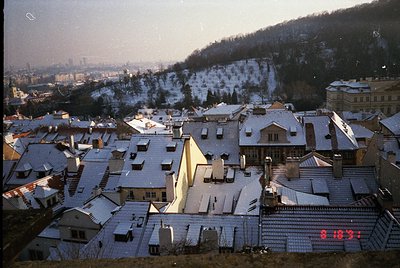 Snow-covered rooftops of a European hillside town, framed by forested hills and distant city skyline. Mid-20th century archit...