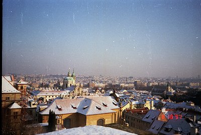 Vintage panoramic shot of Prague’s historic rooftops dusted with snow, showcasing Baroque architecture. Prominent spire and d...