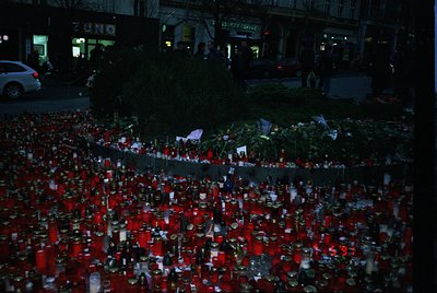 Memorial tribute featuring thousands of red candles and floral arrangements lining a barrier, likely in remembrance of a trag...