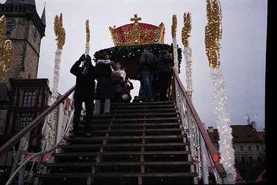 A festive procession ascends a wooden staircase toward a grand, illuminated throne-like structure adorned with a golden crown...