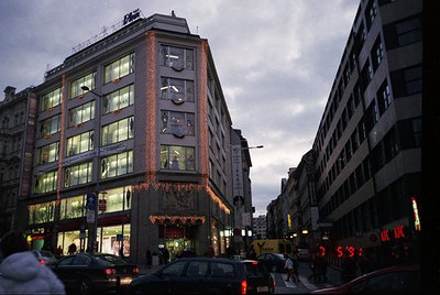 Mid-20th century urban street scene featuring a corner building with illuminated signage and festive garlands. Brick facade w...