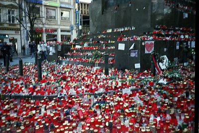Memorial tribute wall covered in red roses, candles, and handwritten notes. Urban setting with buildings and street lamps in ...