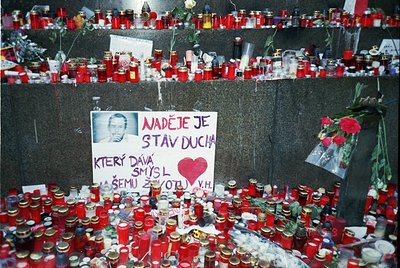 Memorial tribute wall covered in red candles, flowers, and handwritten notes in Czech. Prominent sign reads *"Naděje je stav ...