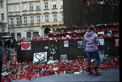 Memorial tribute wall lined with red candles, flowers, and handwritten notes in an urban plaza. A large red heart symbol domi...