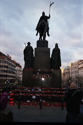 Equestrian statue monument with flanking figures, surrounded by red candles and floral tributes. Urban setting with historic ...