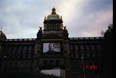 Neoclassical building with domed roof and statues, featuring a large projected portrait of a man on its facade. Likely Prague...