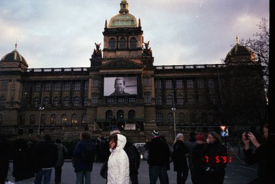 Neoclassical Prague Castle facade with projected portrait of Vaclav Havel, 1989 Velvet Revolution. Crowd gathers below illumi...