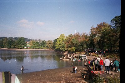 Vintage lakeside gathering with dense foliage framing a calm water body. Group of people in casual 1970s–80s attire—some wadi...