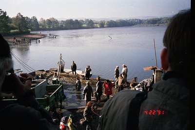 Vintage fishing scene with wooden docks and boats on a calm lake, likely Eastern Europe . Crowd in layered clothing—some hold...