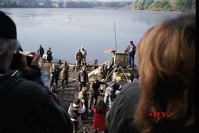 Rustic lakeside scene featuring a wooden dock with a makeshift fishing platform. Crowd of onlookers, including children, gath...