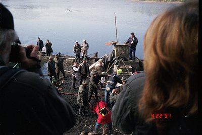 A group of people on a wooden pier by a lake, likely for a ceremonial or public event. Individuals wear 1970s-era clothing (o...